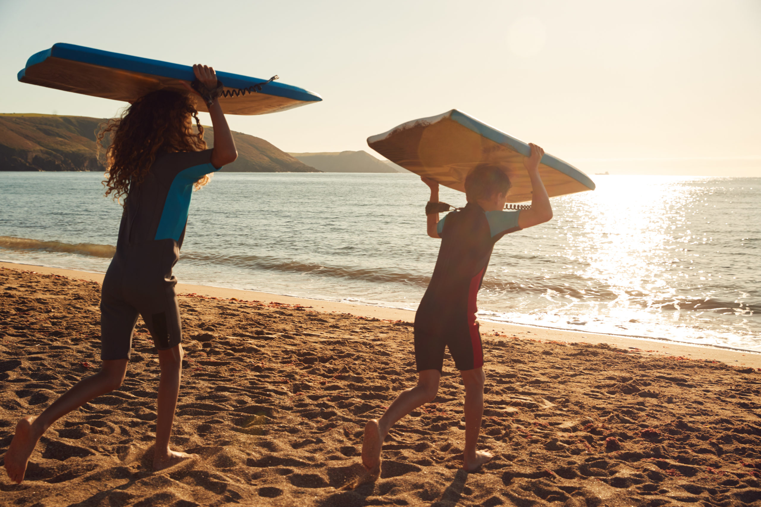 Rear View Of Children In Wetsuits Carrying Bodyboards On Summer Beach Vacation Having Fun By Sea